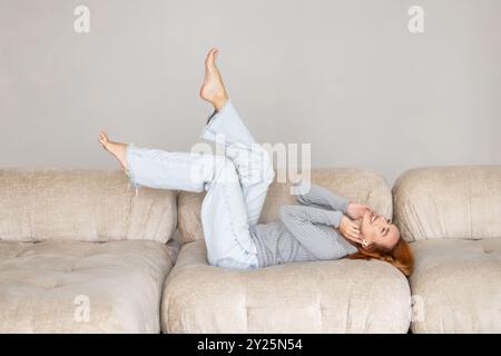 Una giovane e tranquilla donna sdraiata su un comodo divano morbido con gambe sollevate si sente spensierata. Trascorri un fine settimana tranquillo da sola a casa. Giorno libero, niente stress, felice proprietario fe Foto Stock