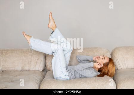 Una giovane e tranquilla donna sdraiata su un comodo divano morbido con gambe sollevate si sente spensierata. Trascorri un fine settimana tranquillo da sola a casa. Giorno libero, niente stress, felice proprietario fe Foto Stock