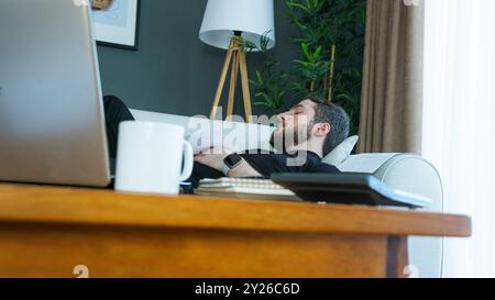 Un uomo con una camicia nera è sdraiato su un divano, mentre fa un pisolino durante una pausa di lavoro. Un portatile, una tazza e dei documenti sono sul tavolo in primo piano, momento di Foto Stock