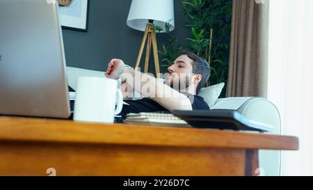 Un uomo riposa su un divano, guardando l'orologio mentre si prende una pausa dal lavoro. Un computer portatile e una tazza da caffè poggiano su un tavolo di legno. Lavoro rilassato da casa r Foto Stock