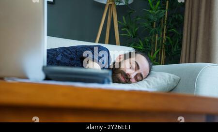 Un uomo sta riposando su un divano, dopo essersi addormentato davanti al suo portatile. Stanchezza o relax dopo una lunga giornata di lavoro o di tempo sullo schermo a casa. Foto Stock