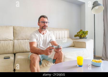 Uomo di mezza età sorridente mentre gioca ai videogiochi sul divano, gustando uno spuntino a casa con la modalità grandangolare Foto Stock