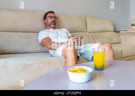 Uomo di mezza età sorridente mentre gioca ai videogiochi sul divano, gustando uno spuntino a casa con la modalità grandangolare Foto Stock