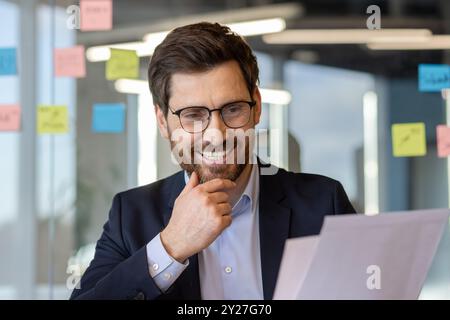 Professionisti che indossano occhiali e sorridono mentre guardano i documenti. Ambiente da ufficio con appunti adesivi su una parete in vetro, che riflette innovazione e produttività. Foto Stock