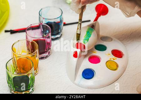 Vista ravvicinata del processo di pittura dell'uovo di Pasqua con pitture e pennelli colorati sul tavolo bianco. Svezia. Foto Stock