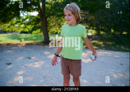 Un bambino di sei anni gioca a petanque in un parco soleggiato, lanciando una palla di metallo. È in piedi con sicurezza, godendo di una divertente attività all'aperto nella natura Foto Stock