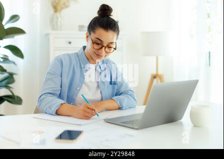 Donna bruna araba o indiana di successo con una camicia blu, seduta a una scrivania mentre lavora sul laptop, svolgendo attività online, sviluppando un nuovo progetto digitale, pianificando una strategia di marketing Foto Stock