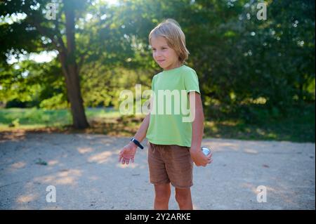 Un bambino di sei anni gioca a petanque in un parco soleggiato, lanciando una palla di metallo. È in piedi con sicurezza, godendo di una divertente attività all'aperto nella natura Foto Stock