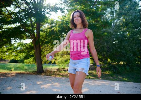 Una donna ama giocare a petanque in una giornata di sole nel parco. Vestita in modo informale con un top rosa e pantaloncini in denim, sorride mentre regge una palla di metallo Foto Stock