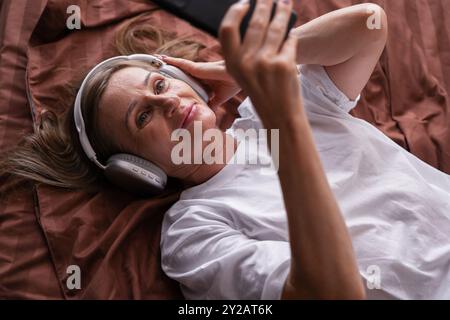 Una donna anziana che utilizza un gadget digitale che naviga su Internet per guardare film o videochiamare online, sdraiata sul letto e svegliata al mattino. Foto Stock