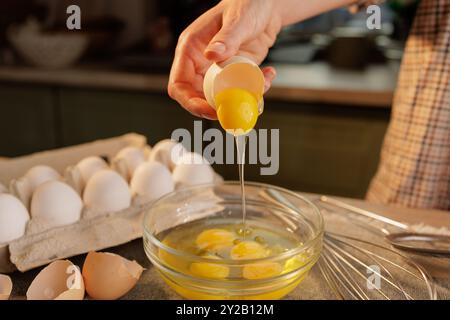 Una persona che spezza un uovo in un recipiente di vetro, con diverse uova in un cartone sullo sfondo. La scena è ambientata in una cucina, che mostra le promesse Foto Stock