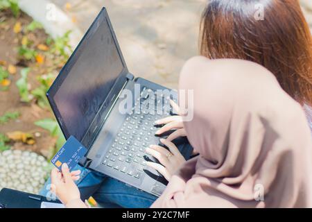 Un paio di donne asiatiche si concentrano su un notebook che discute di shopping online tenendo penne, libri e appunti nel parco del campus. studentesse per ed Foto Stock