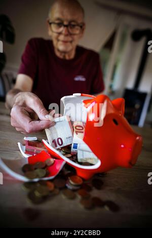 Symbolfoto zum Thema Finanzen im alter - Altersarmut. Ein Alter Mann sitzt zu Hause am Tisch vor einem zerbrochenen Sparschwein und zaehlt Geld. Berli Foto Stock