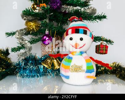 Pupazzo di neve giocattolo a maglia che indossa un cappello e una sciarpa con la scritta dorata "buon Natale!" Vicino a un albero di Natale decorato artificialmente. Foto Stock