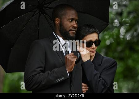Immagine media di una donna anziana che tiene il marito solidale con il braccio in lutto alla cerimonia commemorativa. Coppia multietnica che piange la perdita in un abbraccio confortante sotto l'ombrello nero al cimitero Foto Stock