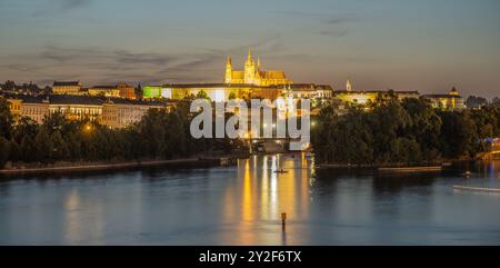 Una vista notturna del Castello di Praga illuminata sul fiume Moldava. Le guglie e gli edifici del castello si riflettono nell'acqua, creando una scena pittoresca. Il paesaggio urbano è immerso nella calda luce dorata, creando un senso di magia e meraviglia. Foto Stock