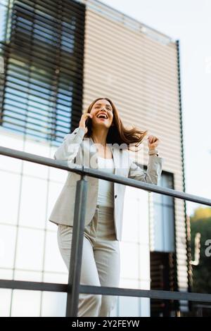 Una donna d'affari gioiosa festeggia un momento all'aperto, partecipando a una allegra conversazione telefonica mentre si appoggia a una ringhiera accanto a un elegante buildi Foto Stock