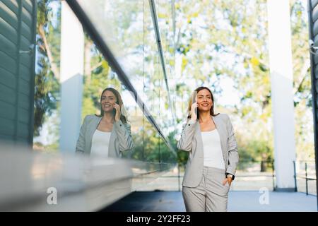 Una donna professionista cammina con facilità, sorridendo mentre si concentra sul telefono, mescolando lo stile di vita moderno con la tranquillità della natura sullo sfondo Foto Stock