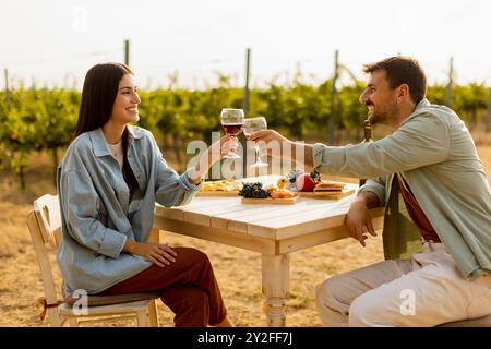 La coppia si rilassa ad un tavolo rustico in un vigneto illuminato dal sole, gustando vino e spuntini gourmet. Condividono risate e gioia, circondati da vigne lussureggianti e il Foto Stock