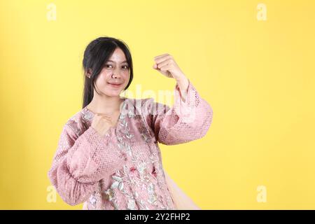 Eccitata donna indonesiana che indossa Kebaya in piedi con le mani strette posa isolata su sfondo giallo Foto Stock