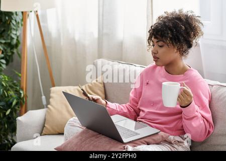 Una giovane donna si rilassa a casa, sorseggiando un caffè e concentrandosi sul suo notebook. Foto Stock