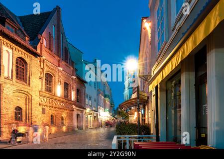 Strada acciottolata della città vecchia illuminata da caldi lampioni di strada la sera. Edifici storici, caffetterie accoglienti e gente sfocata dal movimento. Freddo destin Foto Stock