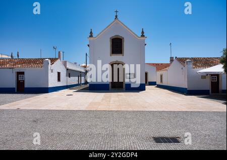Cappella imbiancata con finiture blu in una tranquilla piazza acciottolata nel villaggio di Porto Covo, Alentejo, Foto Stock