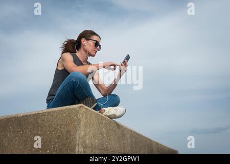 Donna seduta che usa il telefono cellulare e indossa auricolari all'aperto Foto Stock