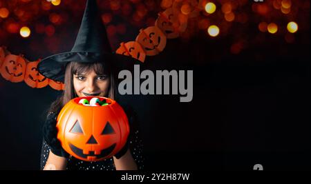 Giovane e bella strega di Halloween che mostra la tazza di Jack o lanterna piena di caramelle con una zucca di carta garland e le luci dietro di lei. Ripresa in studio. Foto Stock