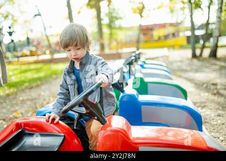Simpatico bambino che cavalca un'auto giocattolo divertendosi in un parco giochi all'aperto nella calda giornata autunnale. Divertimento attivo per i bambini in autunno. Foto Stock