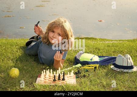 Kid mostra pezzi di scacchi su una scacchiera. Sviluppo iniziale. Ragazzo che pensa agli scacchi nel parco estivo. Il concetto di apprendimento e di crescita dei bambini Foto Stock