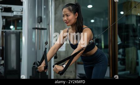 Una donna asiatica sportiva e in forma si sta allenando su una macchina multi-stazione in palestra, concentrandosi sull'allenamento delle spalle e delle braccia. persone a Foto Stock