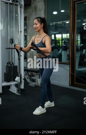 Una donna asiatica sportiva e in forma si sta allenando su una macchina multi-stazione in palestra, concentrandosi sui bicipiti, in piedi e tirando la cabl Foto Stock