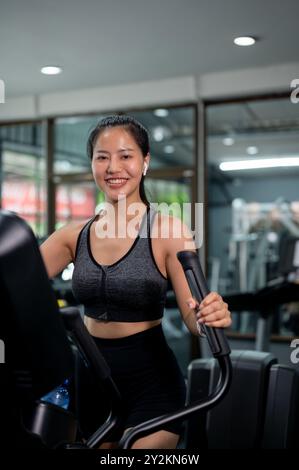 Una bella e sportiva donna asiatica in abbigliamento sportivo corre con una macchina ellittica da cross trainer in palestra, sorridendo alla telecamera. allenamento cardio, sforzo Foto Stock