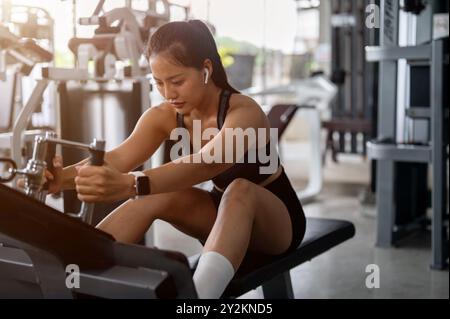 Una bella e sportiva donna asiatica in abbigliamento atletico è seduta su una panchina, tirando un cavo per allenare i muscoli della schiena, allenandosi su una stazione multipla Foto Stock