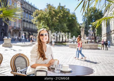 Una donna siede ad un tavolo da caffe' all'aperto, sorridendo mentre sorseggia un drink. La luce del sole filtra tra tra gli alberi in una bellissima piazza piena di gente enjo Foto Stock