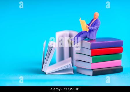 Concetto di apprendimento e conoscenza con un uomo in miniatura che indossa una tuta viola, si siede su libri vivaci e legge da una cartella aperta. Foto Stock