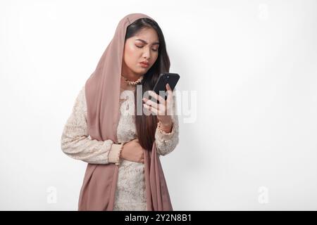 Giovane donna musulmana asiatica in velo hijab ha fame mentre tiene lo stomaco e usa uno smartphone per ordinare cibo. Ramadan e Eid Mubarak Foto Stock