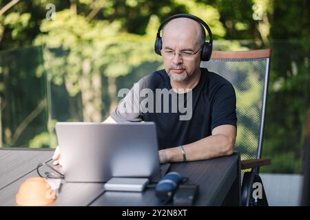 Un uomo maturo che lavora in remoto da casa indossando cuffie e occhiali. Uomo Foto Stock