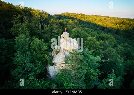 Veduta aerea di un alpinista in cima alla formazione rocciosa di Dovbush Rocks nelle montagne dei Carpazi, Ucraina. Il sole tramonta, proiettando un caldo bagliore sulla lussureggiante foresta verde, sulle colline lontane, creando una vista mozzafiato. Foto Stock