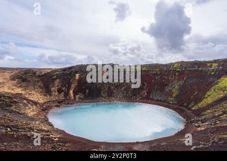 Una vista serena del cratere Kerid, una caldera vulcanica in Islanda. Il profondo lago blu-verde contrasta splendidamente con la roccia vulcanica rossa e il verde lussureggiante Foto Stock