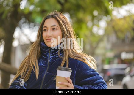 Ragazza latina che beve un caffè seduto su una panchina in un parco pubblico. Foto Stock