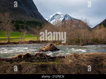 Il tranquillo fiume si snoda attraverso una valle verde, circondata da alberi senza fronzoli e torreggianti montagne ricoperte di neve, mentre le nuvole serali si radunano Foto Stock