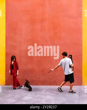 Una giovane coppia gode di una tranquilla passeggiata con le loro distese lungo una strada vivace e colorata, la donna vestita di rosso e l'uomo vestito in modo informale Foto Stock
