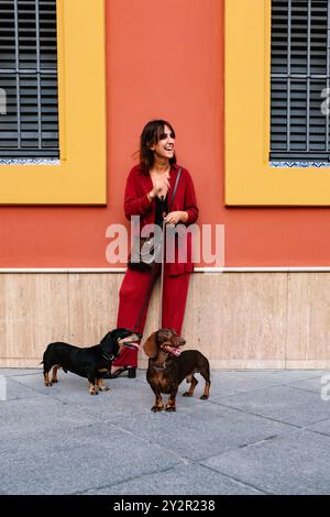 Una giovane donna sorridente in un vivace vestito rosso porta con gioia i suoi due cani Dachshund su un sentiero illuminato dal sole con uno sfondo colorato Foto Stock