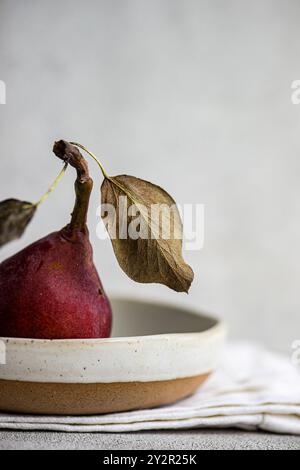 Una pera organica matura, con una pelle rossa vivace e delicate foglie secche attaccate al gambo, si trova elegantemente in una ciotola di ceramica dai colori terrosi Foto Stock