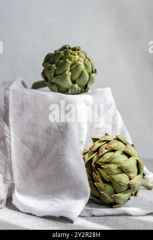 Due carciofi freschi e verdi poggiano su un panno di lino delicatamente drappeggiato, che mostra la loro texture dettagliata e naturale con uno sfondo morbido e neutro Foto Stock
