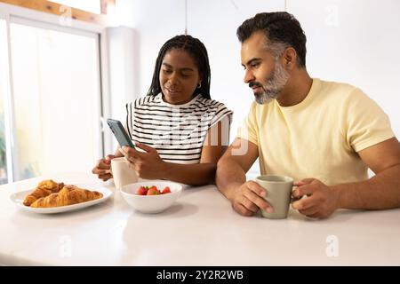 Una allegra donna nera e un uomo indiano si siedono insieme al bancone della cucina, gustando una colazione a base di croissant e frutta mentre si utilizza uno smartphone Foto Stock