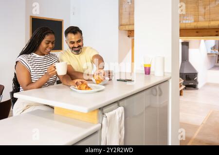 Una coppia di mezza età condivide una deliziosa mattinata sorseggiando un caffè e gustando croissant in una cucina moderna e luminosa Foto Stock