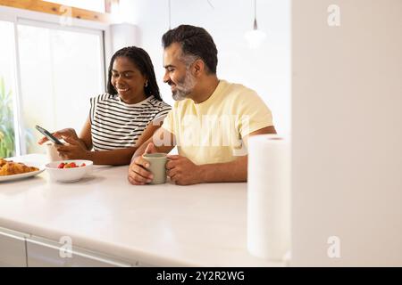 Una coppia multirazziale condivide una mattinata gioiosa in una luminosa cucina, dove si gusta una colazione informale mentre si divertono con uno smartphone Foto Stock